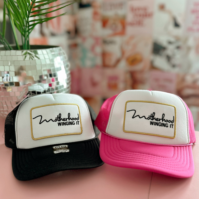 Black-and-white and pink-and-white “Motherhood Winging It” trucker hats displayed together.