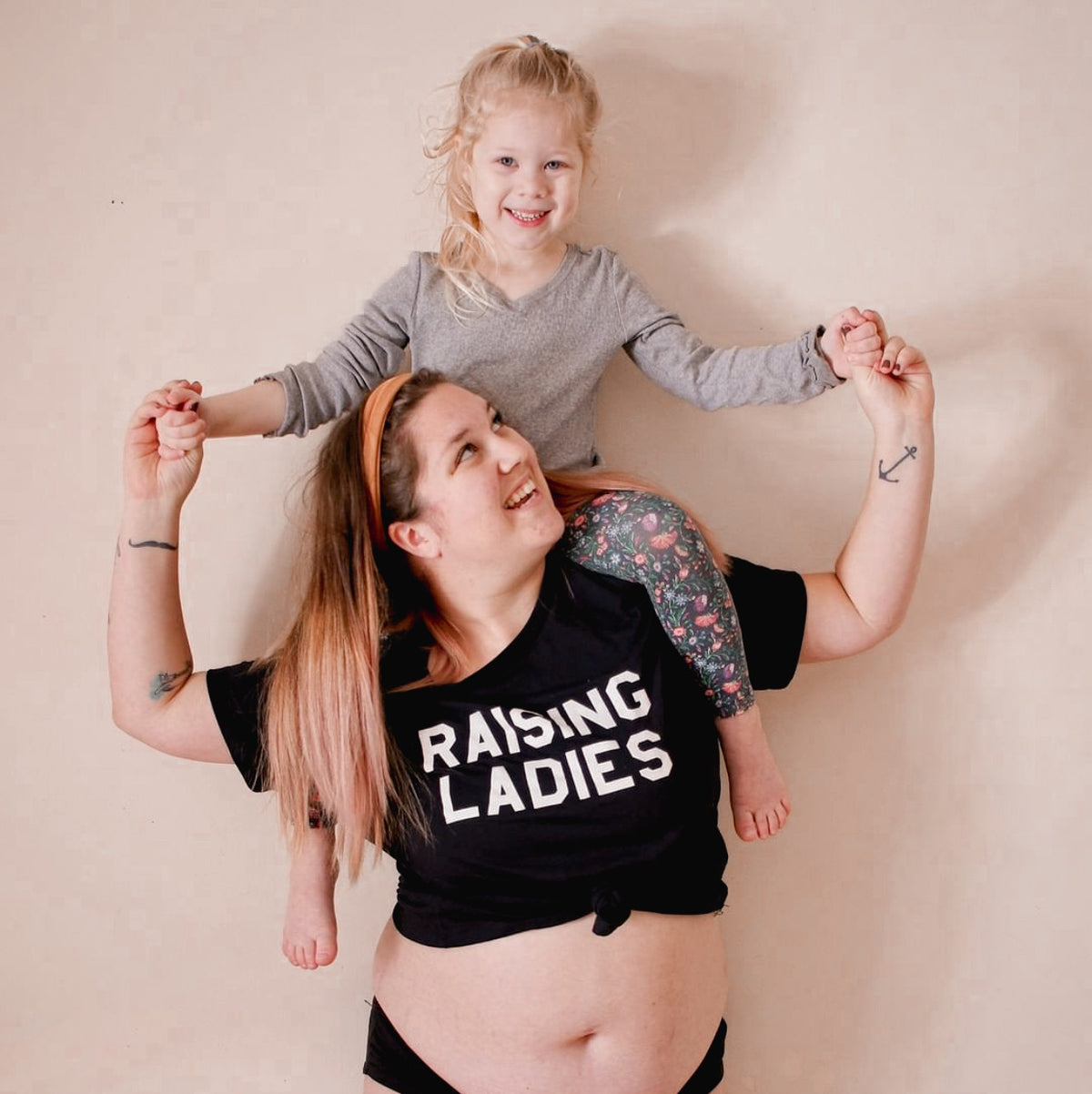 Woman wearing a black “Raising Ladies” T-shirt with a child sitting on her shoulders.