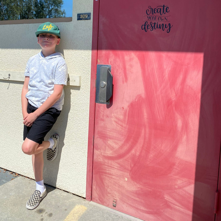 Child in a green cap standing beside a pink school door.