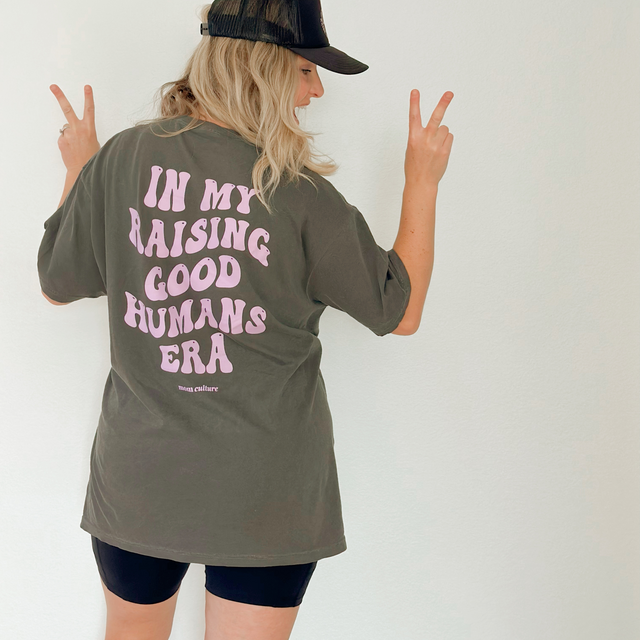Woman wearing an “In My Raising Good Humans Era” T-shirt and holding up two peace signs.