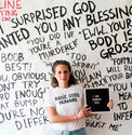 Woman wearing a cream “Raise Good Humans” T-shirt in front of a wall covered in handwritten phrases.