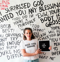 Woman wearing a cream “Raise Good Humans” T-shirt in front of a wall covered in handwritten phrases.