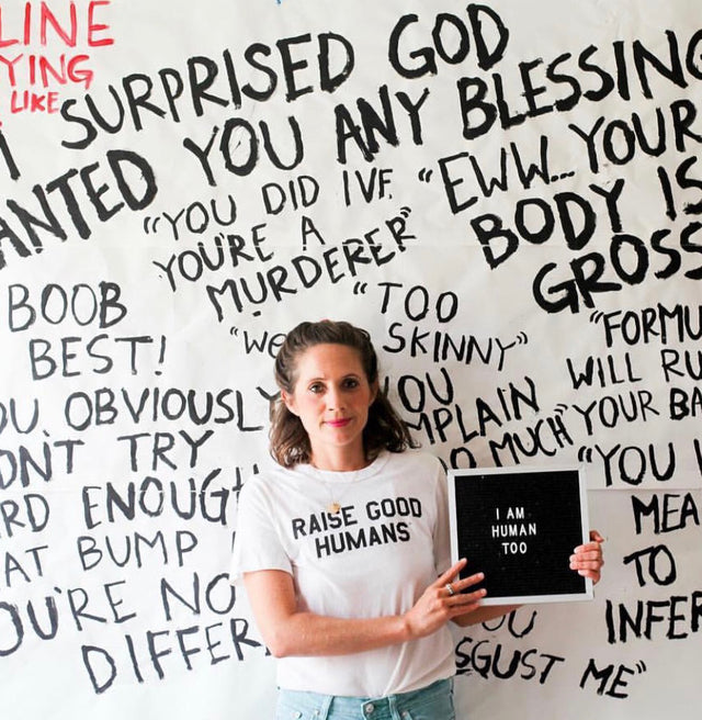 Woman wearing a cream “Raise Good Humans” T-shirt in front of a wall covered in handwritten phrases.