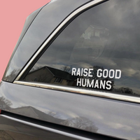 White “Raise Good Humans” decal on a car’s rear side window.