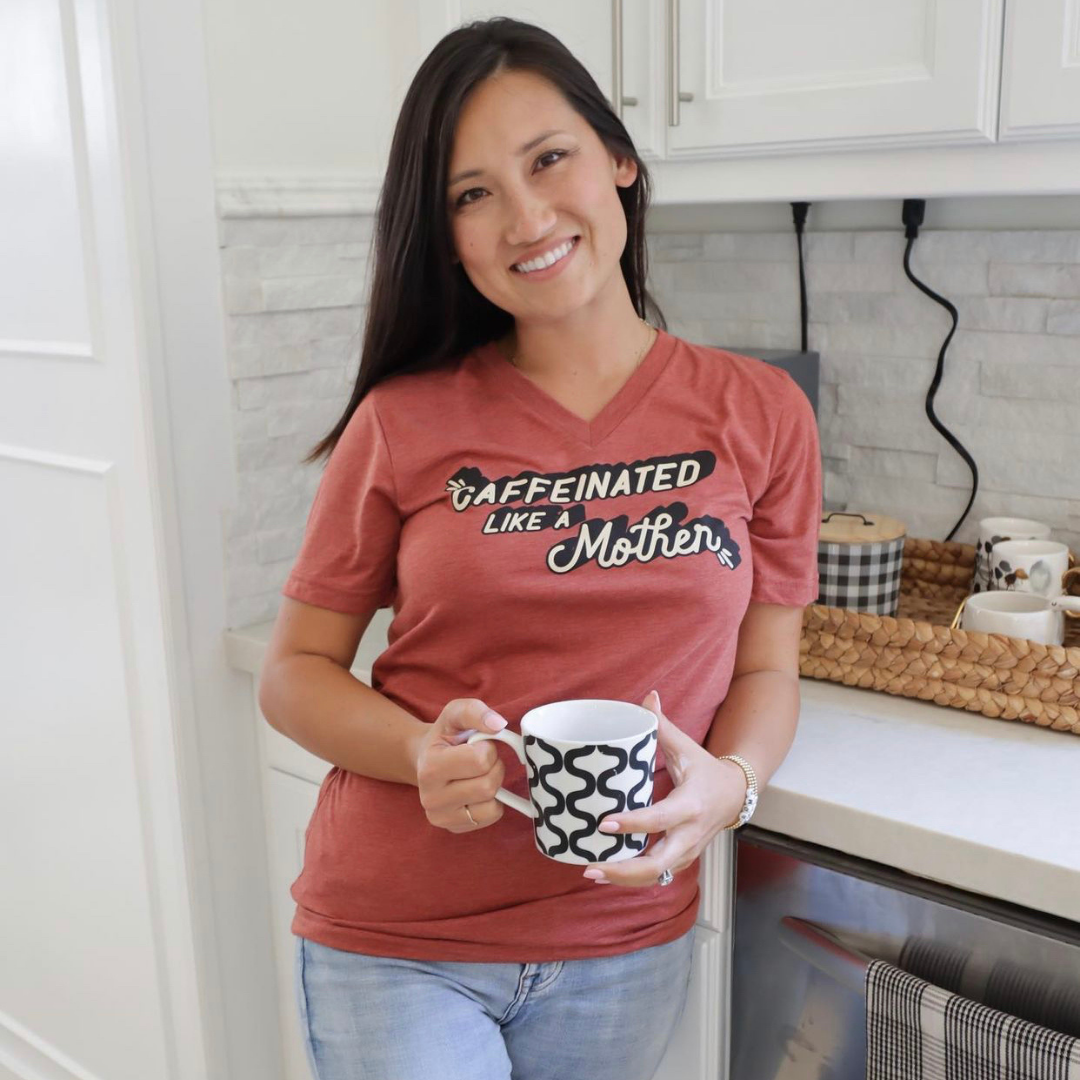 Woman holding a mug while wearing a rust-colored “Caffeinated Like a Mother” V-neck T-shirt.