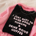 Folded black T-shirt reading “I Just Want to Raise Good Humans & Drink Good Coffee” on a pink blanket.