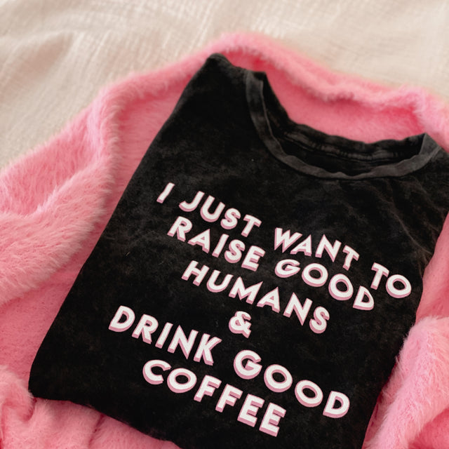 Folded black T-shirt reading “I Just Want to Raise Good Humans & Drink Good Coffee” on a pink blanket.