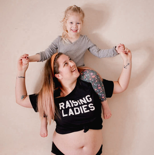 Woman wearing a black “Raising Ladies” T-shirt with a child sitting on her shoulders.