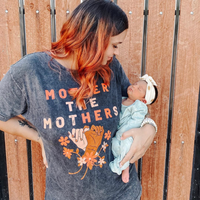 Woman holding a baby while wearing a dark gray “Mother the Mothers” T-shirt with an orange floral design.