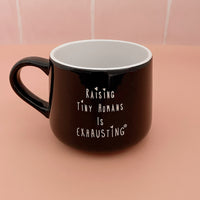 Black ceramic mug with a white interior and “Raising Tiny Humans Is Exhausting” lettering.
