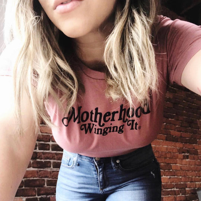 Woman wearing a dusty pink “Motherhood Winging It” T-shirt with jeans.