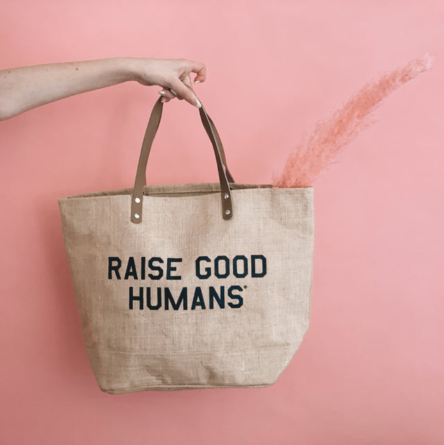 Natural jute tote with brown handles and black “Raise Good Humans” lettering.